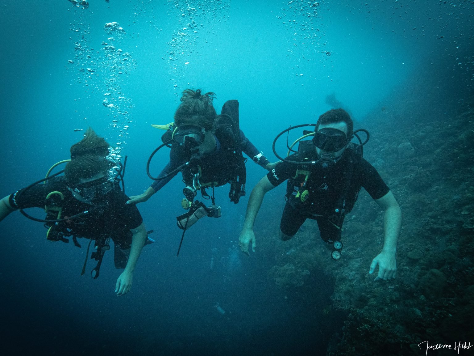 Wide-angle photograph by Maxime Hiblot, captured at the USAT Liberty wreck dive site in Amed, Bali, featuring a PADI instructor ensuring the safety of two beginner divers during a Discover Scuba Diving (DSD) experience. The instructor's professionalism and the awe of the novice divers are palpable.